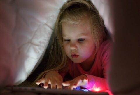 Little girl is playing on the tablet in secret from her parents at night under the blanket.
