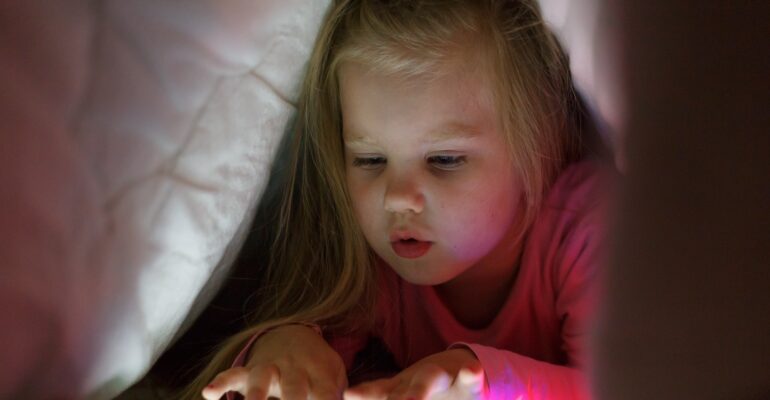 Little girl is playing on the tablet in secret from her parents at night under the blanket.